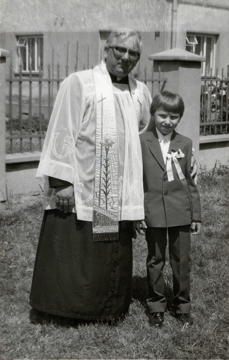 Pan Bazan pozuje wraz z ks. S. Ujdą do pamiątkowego zdjęcia z okazji I Komunii Św. na tle budynku plebanii w Głogowie.