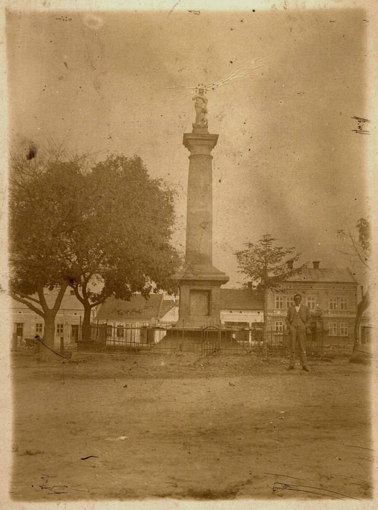 Mężczyzna pozuje do zdjęcia obok figury Matki Boskiej na Rynku w Głogowie. W tle widoczny jest wóz zaprzęgowy i kamienice północnej pierzei Rynku od numeru 19 do 23.