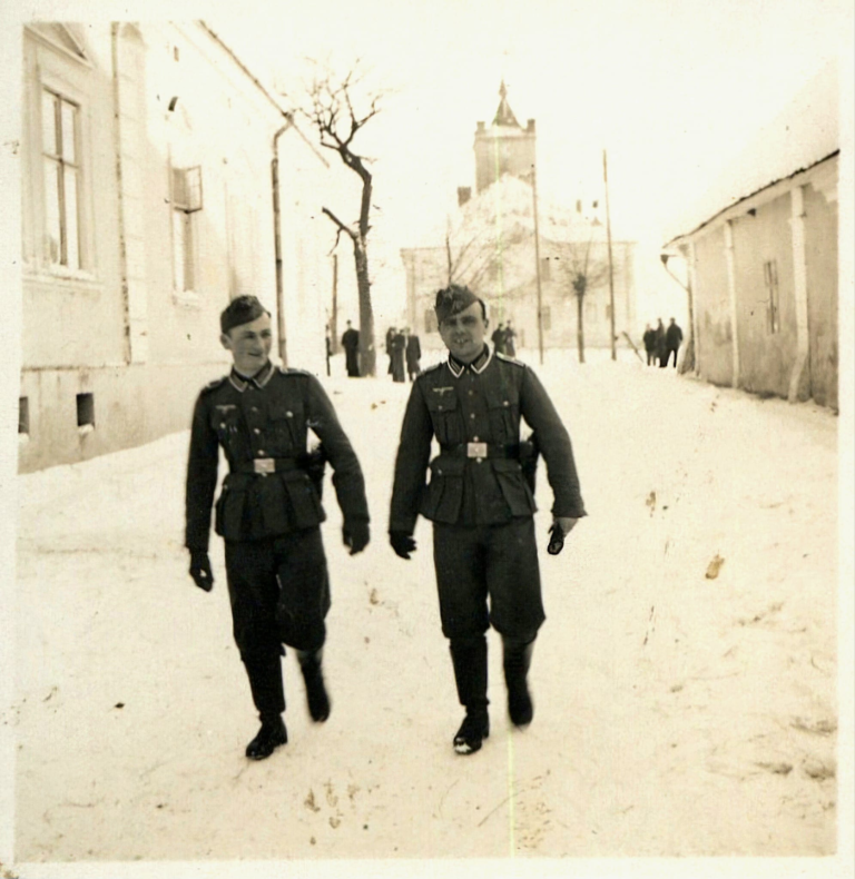 Dwóch niemieckich żołnierzy idzie po śniegu ulicą Sikorskiego w Głogowie. Z tyłu za nimi widać grupę ludzi. W tle od lewej widoczna jest kamienica nr 19 w Rynku , ratusz miejski i kamienica nr 18.