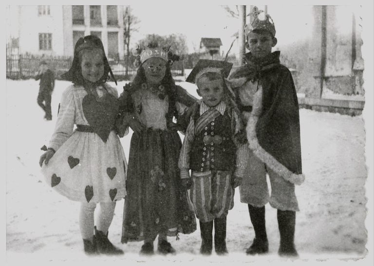 Czworo dzieci w ubranych w różne stroje (kolędnicy , I dzień wiosny ?) pozuje do zdjęcia na podwórku szkolnym , na tle domu nauczyciela i parku „Grabina”.