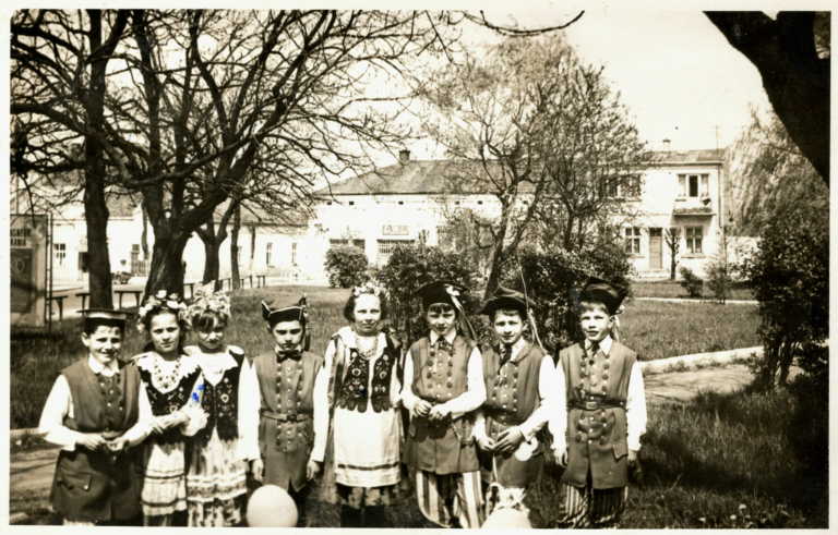 Grupa dzieci w strojach ludowych pozuje do zdjęcia na plantach w Głogowie nieopodal figury Matki Boskiej. W tle widoczne są kamienice w Rynku od numeru 17 do numeru 21.