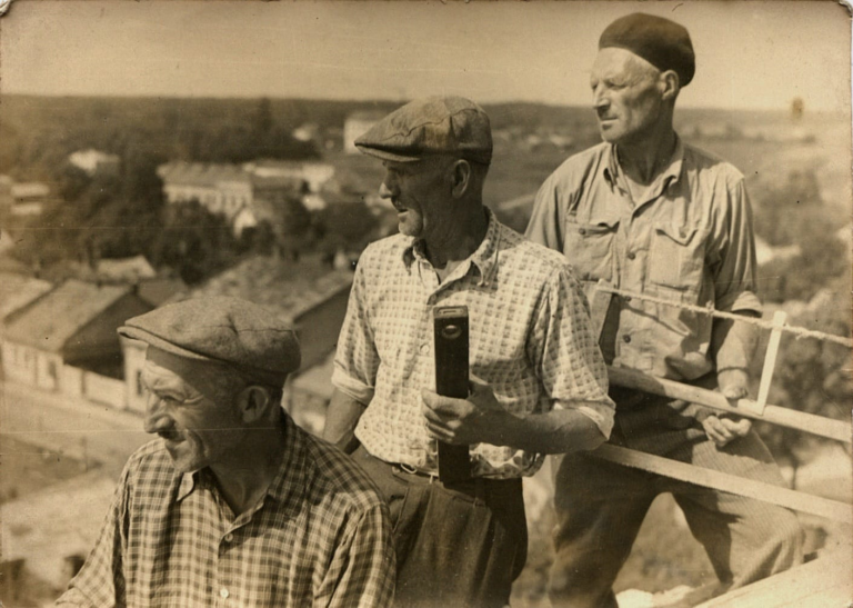 Trzech cieśli stoi na drewnianej konstrukcji wieży zegarowej ratusza w Głogowie podczas jego przebudowy. W tle widoczna północno – zachodnia część Głogowa.