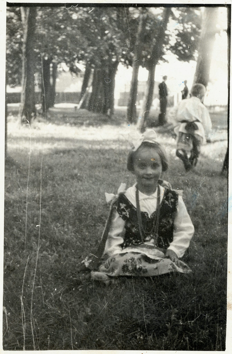 Pani Zofia Brudek (z d. Bazan) w stroju ludowym pozuje do zdjęcia w parku miejskim „Grabina” w Głogowie. W tle widać kilka osób , jedną z nich jest p. Kazimierz Hul.