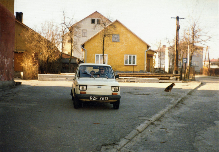 Pani Anna Bąk z mężem i dzieckiem siedzą w zaparkowanym Fiacie 126p obok budynku dzisiejszego liceum. W tle widać kilka budynków przy ulicy Krzywej : nr 1 , nr 3 , nr 12 i nr 16.